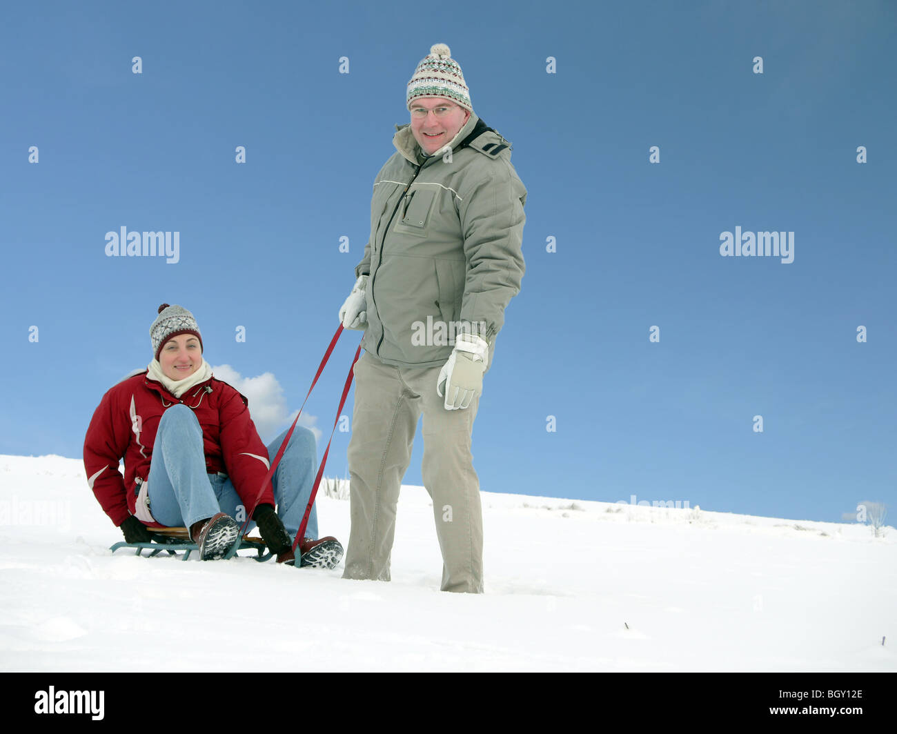Junger Mann zerrte seine Frau sitzen auf Schlitten im Schnee über blauen Himmel Stockfoto