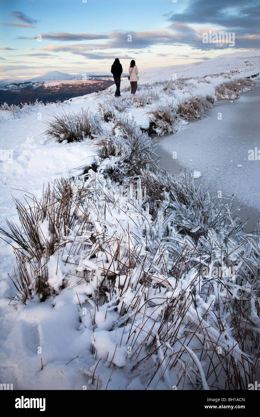 Paare, die im Schnee auf Mauren neben gefrorenen Keeper Teich und Zuckerhut in Entfernung Blorenge Mountain Wales UK Stockfoto