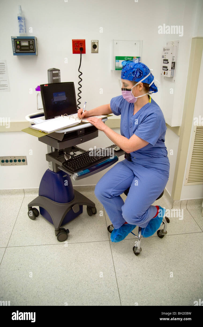 In "Scrubs" gekleidet und mit Hilfe eines Computers für einen Schreibtisch, füllt eine Frau Chirurg rechtlich erforderlichen Unterlagen in einem OP-Saal. Stockfoto