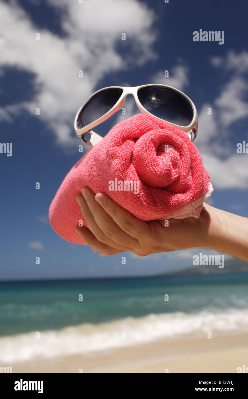 Die Hand eines Mannes hält eine gerollte rote Badetuch und eine weiße Sonnenbrille an einem tropischen Strand Stockfoto