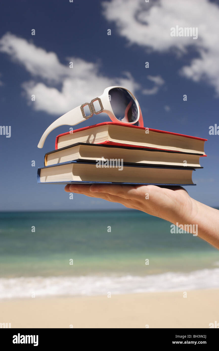 Die Hand eines Mannes hält drei gebundene Bücher und eine weiße Sonnenbrille an einem tropischen Strand Stockfoto