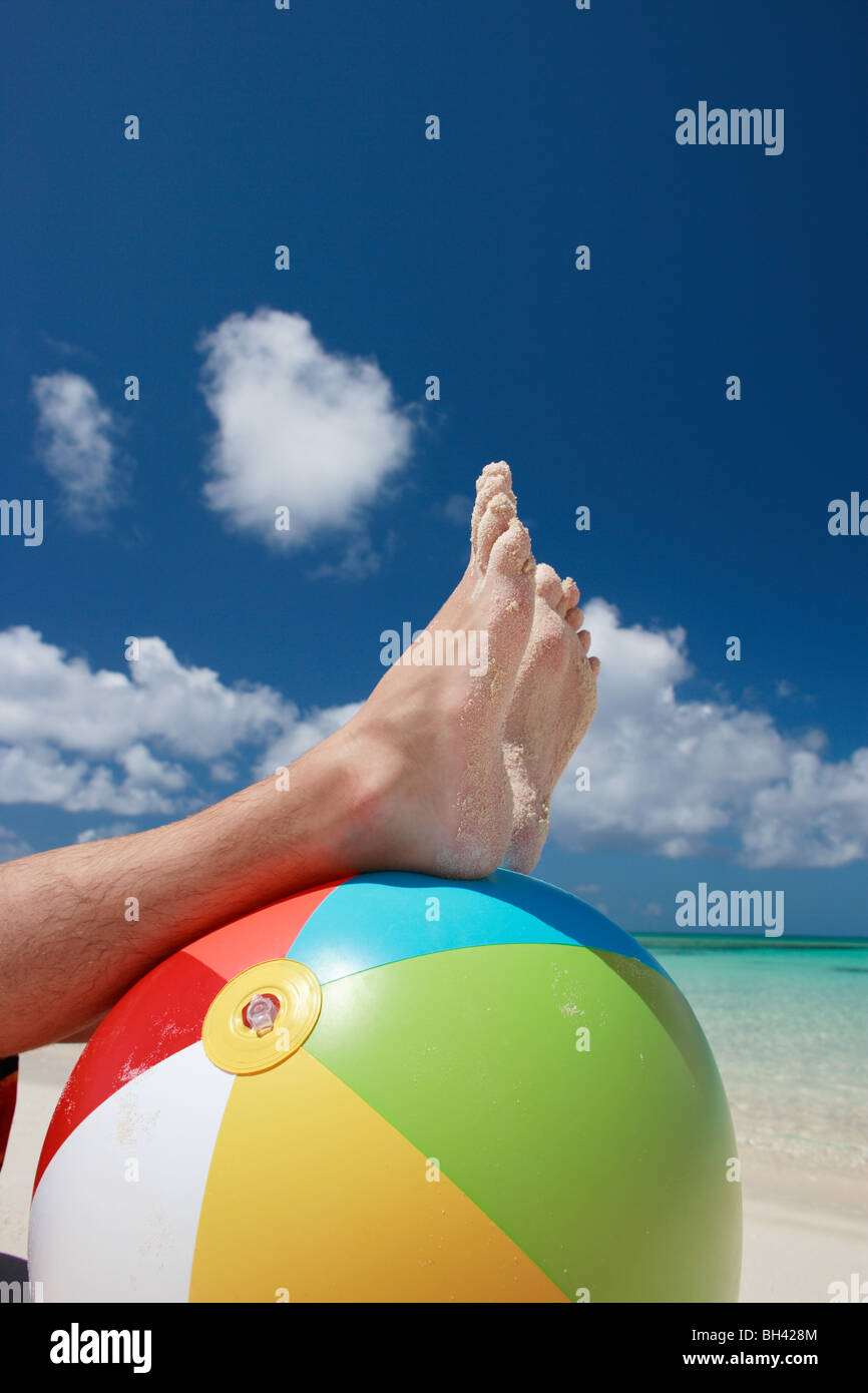 Ein Mann Beine und Füße auf eine aufblasbare Multi farbige Beach-Ball an einem tropischen Strand Stockfoto