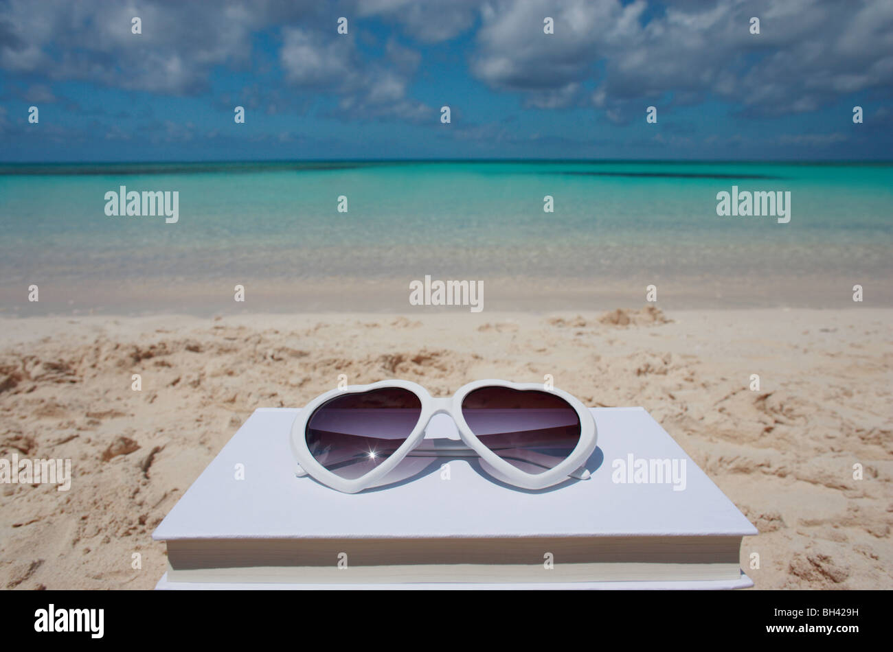 Ein paar weiße herzförmige Sonnenbrille ruht auf einem Buch auf einem einsamen tropischen Strand Stockfoto