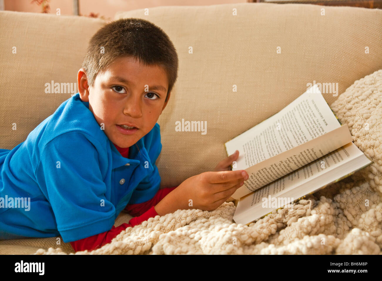 Hispanic 8-9 Jahre alter Junge mit einem Buch auf dem Sofa. United States, Augenkontakt, regnerischen Tag Aktivität POV HERR United States Stockfoto