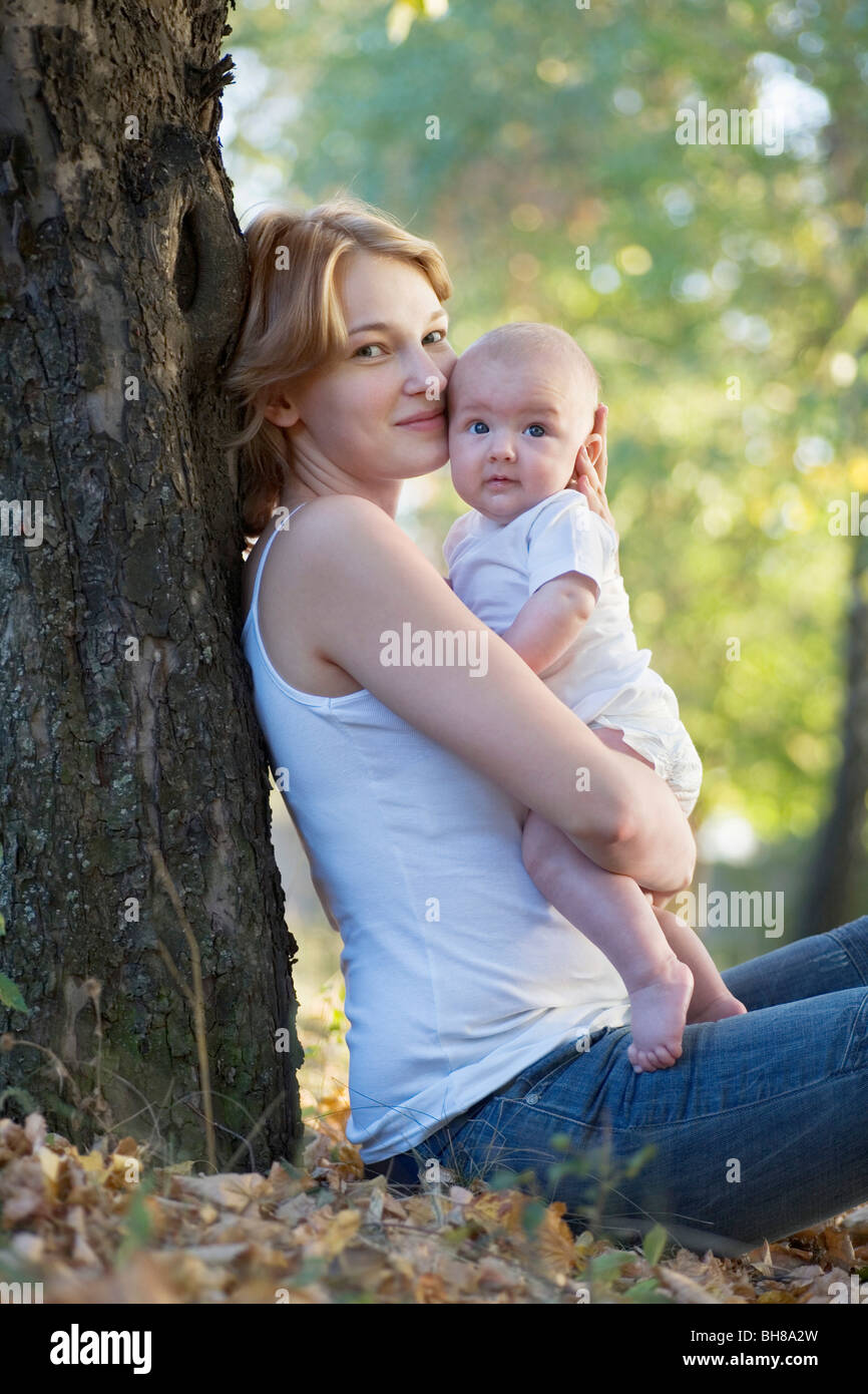 Eine Mutter sitzen gegen einen Baum, küssen ihr Baby, Blick in die Kamera Stockfoto