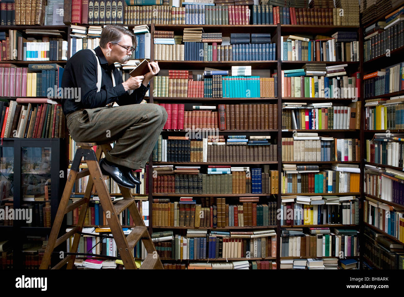 Ein Mann sitzt auf einer Leiter in einer Buchhandlung ein Buch zu lesen Stockfoto