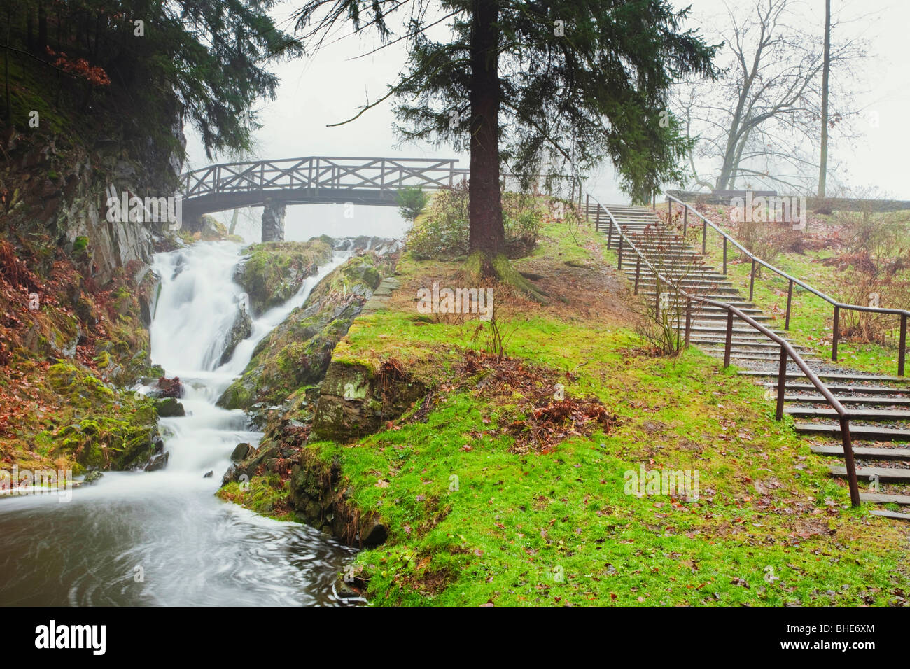 Ruhiger Wasserfall, der unter einer hölzernen Brücke in einem üppigen Park der Tschechischen Republik fließt, mit Stufen, die auf einen grasbewachsenen Hügel führen Stockfoto