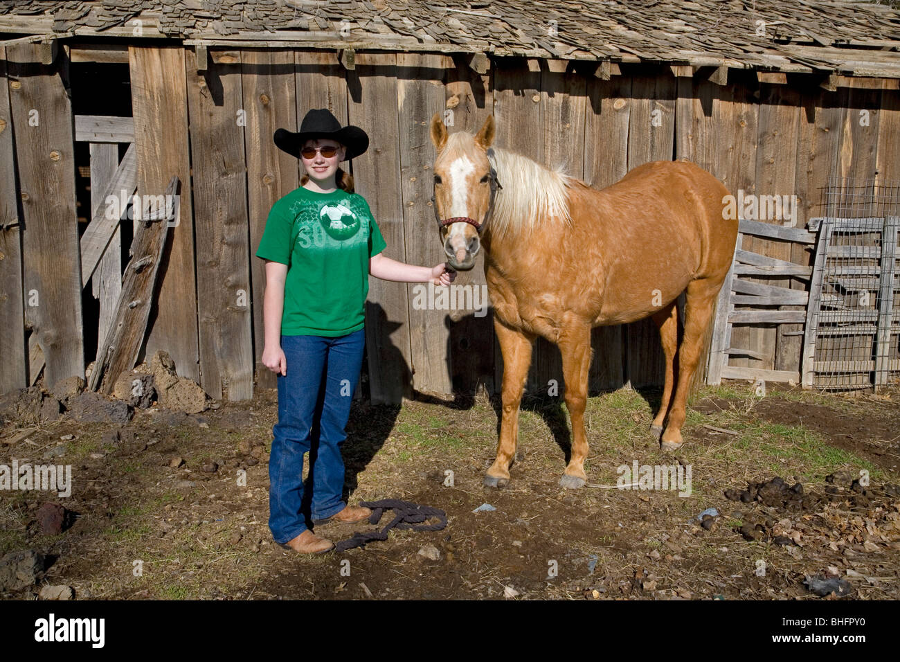 Ein junges Cowgirl mit einem zehn Gallonen-Hut und ihrem Palomino-Pferd vor einer alten Scheune. Stockfoto