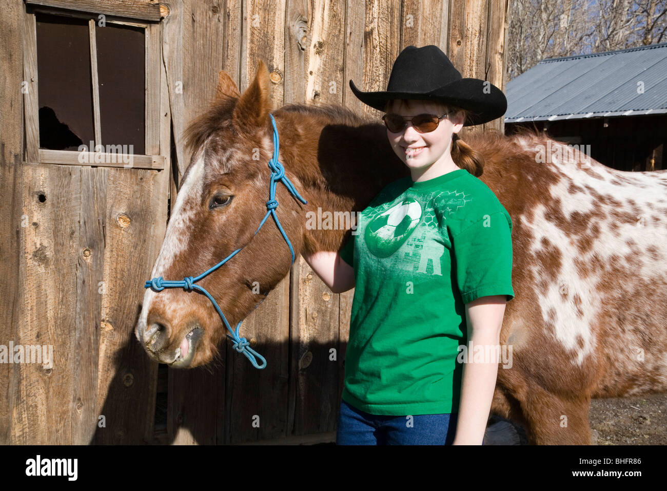 Ein junges Cowgirl mit einem zehn Gallonen-Hut und roan Appaloosa Pferd vor einer alten Scheune. Stockfoto