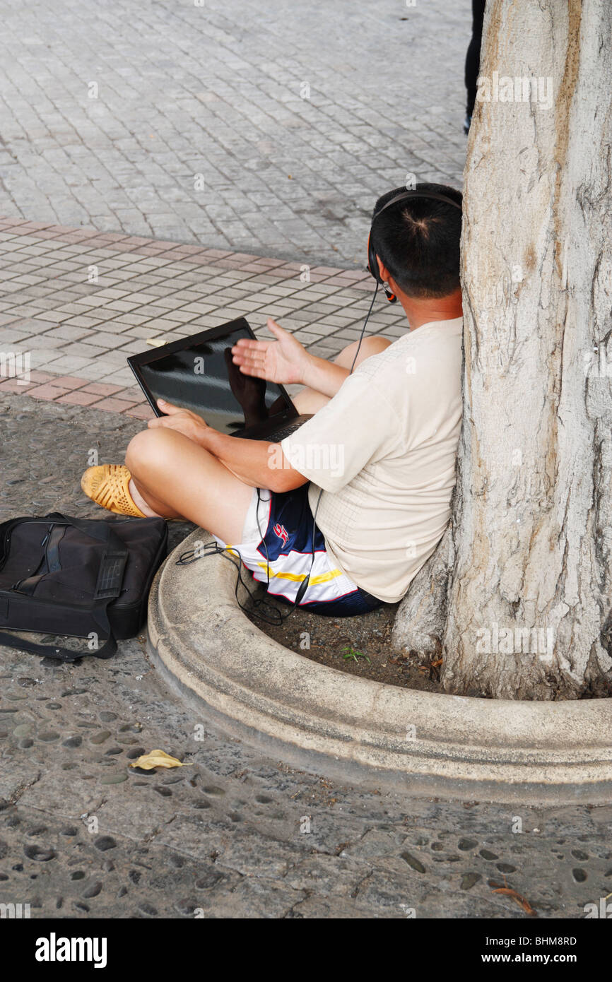 Chinesischer Mann mit Laptop und Kopfhörer sitzen unter Baum in WiFi-Zone. Stockfoto