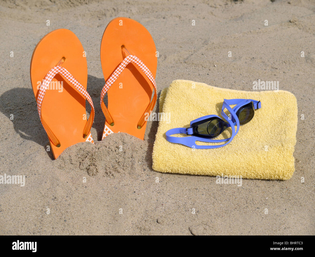 Orange Flip Flops, die vertikal in Strandsand und Schwimmbrille auf gelben Badehandtuch getrieben Stockfoto