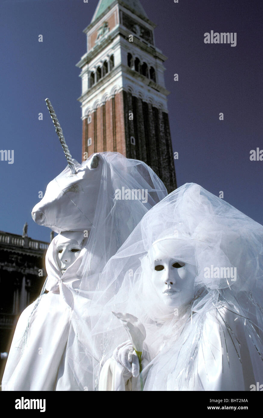 Paar in weißen Kostümen, mit Einhorn Hut, andere halten Calla Lily, San Marco Campanile hinter, Karneval in Venedig, Italien Stockfoto