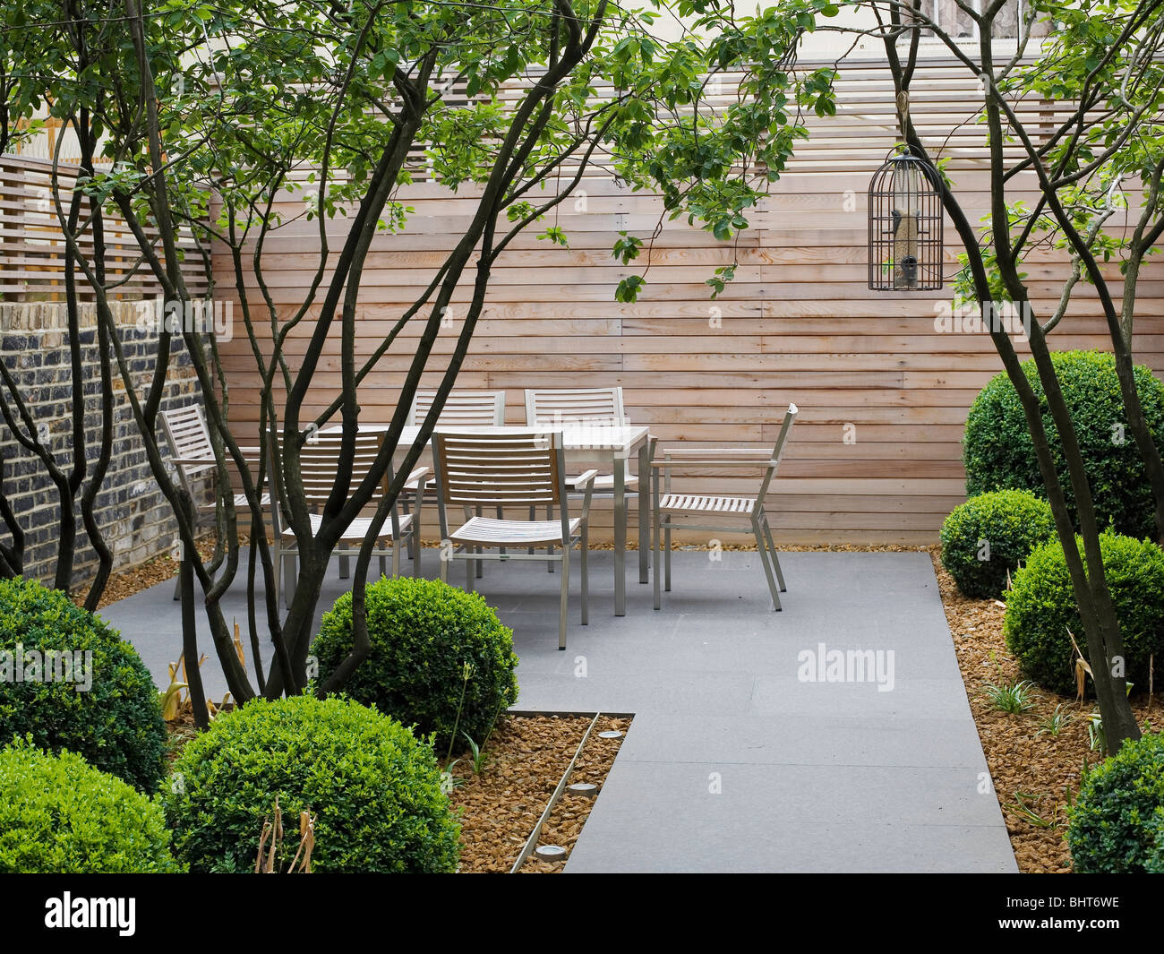 Moderner Tisch und Stühle gegen hohen Holzzaun in architektonischen Reihenhaus Garten mit beschnittene Box Sträucher und graue Pflaster Stockfoto
