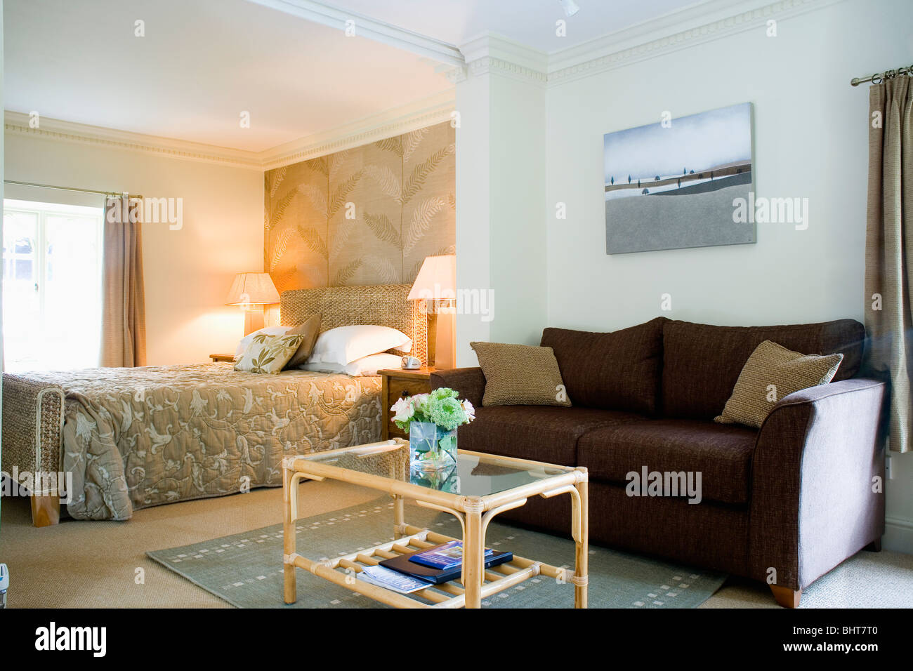 Zuckerrohr und Glas Tisch vor braunen Sofa im Stadthaus Schlafzimmer mit brennenden Lampen auf beiden Seiten des Bettes mit Beige Bettdecke Stockfoto