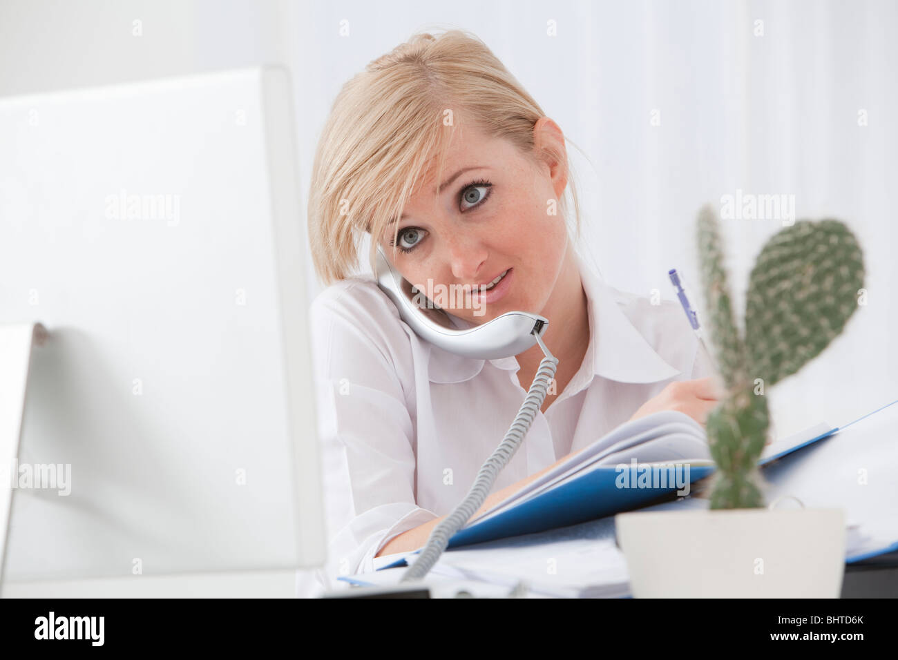 junge Frau beschäftigt Multitasking vor Computer-Bildschirm und am Telefon an ihrem Schreibtisch in einem klaren, modernen Büro Stockfoto
