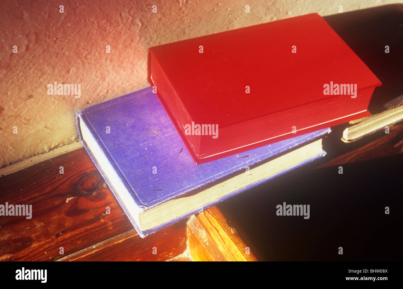 Eine rote und eine blaue Buch auf hölzernen Sims in warmen Abend Sonnenlicht und werfen lange Schatten auf strukturierte Creme Wand sitzen Stockfoto