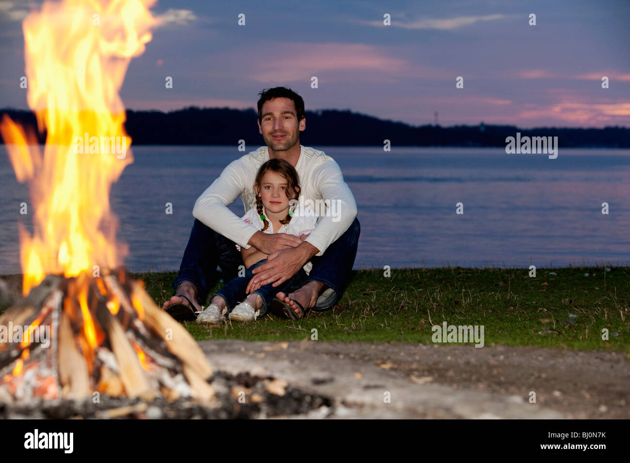 junge Mädchen und Vater bei Sonnenuntergang am Lagerfeuer sitzen Stockfoto