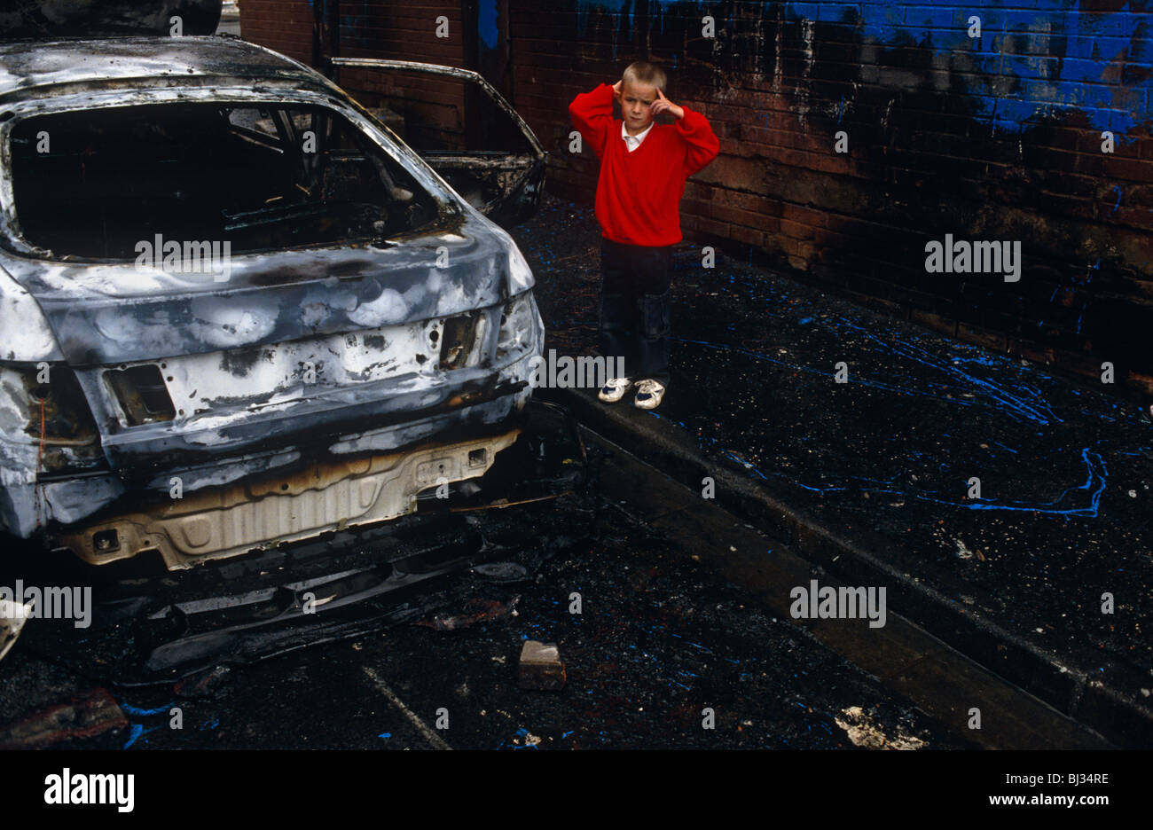 Ein kleiner Junge sieht traumatisierten stand neben einer ausgebrannten Shell eine Limousine von Vandalen in Belfast in Brand gesteckt. Stockfoto