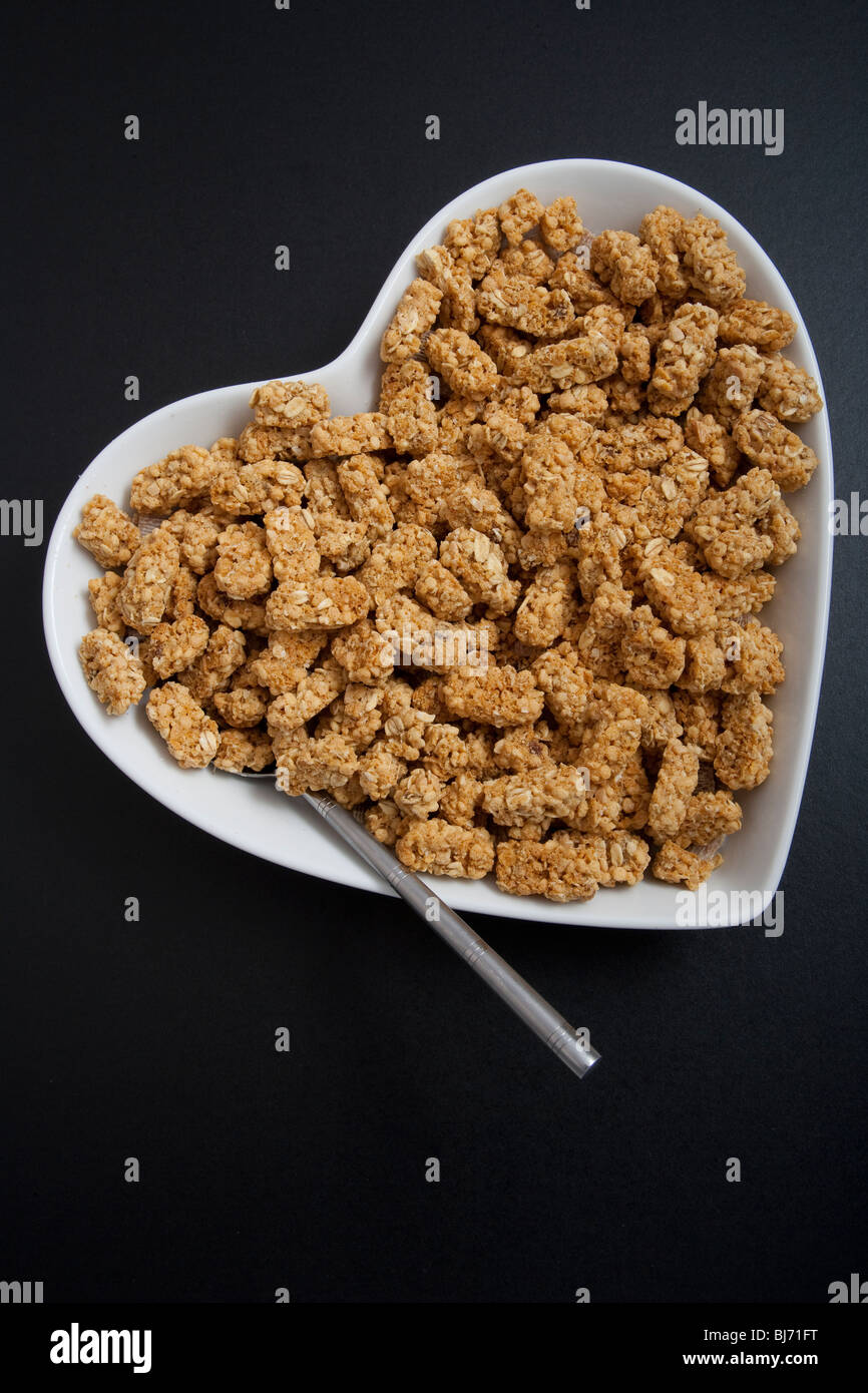 Herzförmige Müslischale oder Schale mit Löffel und Getreide auf schwarzem Hintergrund. Stockfoto