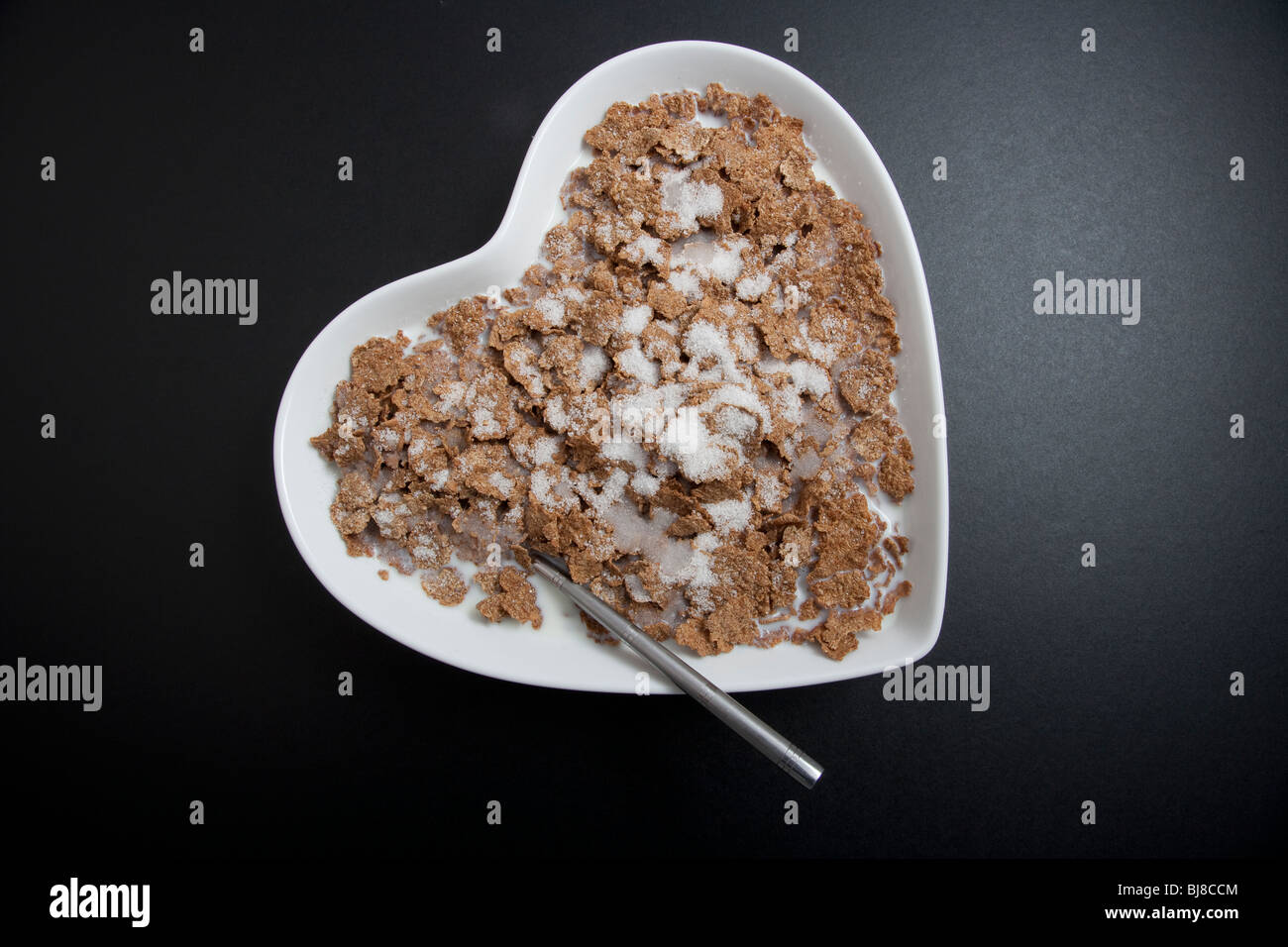 Herzförmige Müslischale oder Schale mit Löffel und Getreide auf schwarzem Hintergrund. Stockfoto