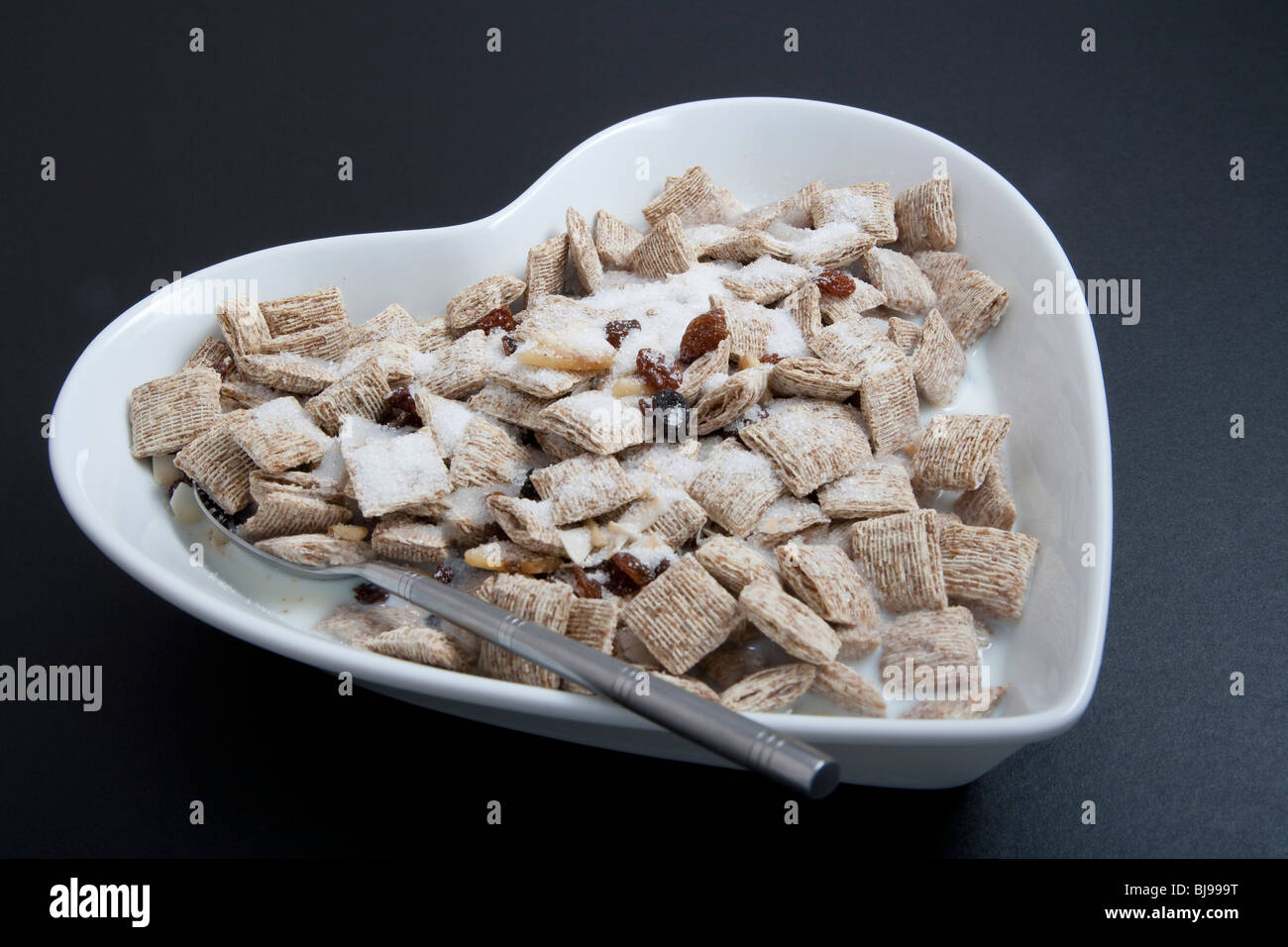 Herzförmige Müslischale oder Schale mit Löffel und Getreide auf schwarzem Hintergrund. Stockfoto