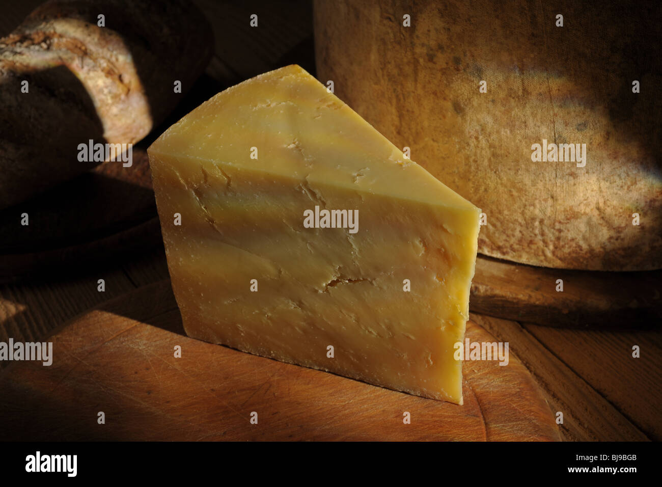Keil reifer Cheddar-Käse an Bord mit ganzen Käse und Brot im Hintergrund Stockfoto