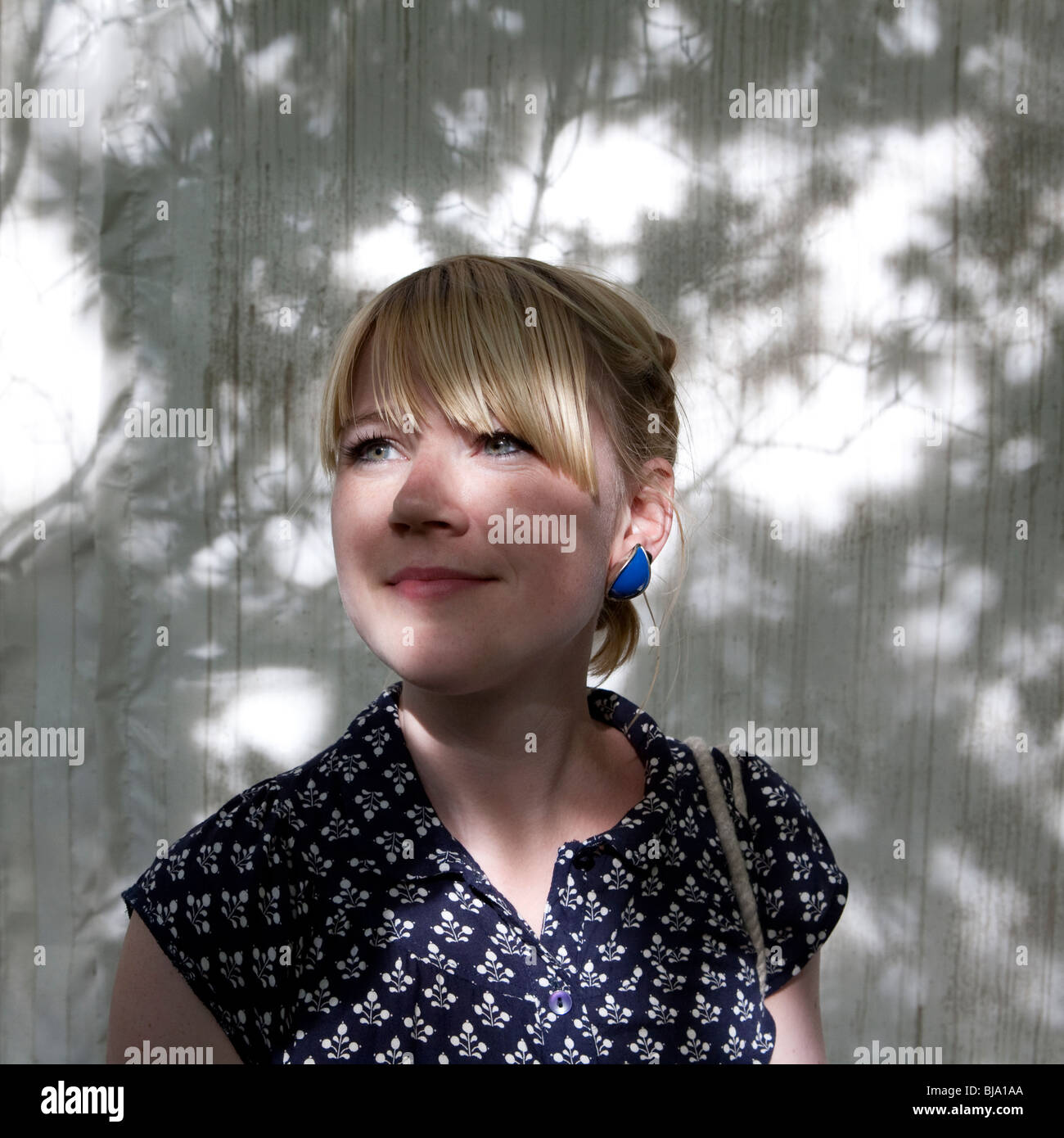 Porträt der jungen blonden Frau mit Fransen und gemusterten Kleid posiert vor der weißen Wand bedeckt, im Schatten der Blätter Stockfoto