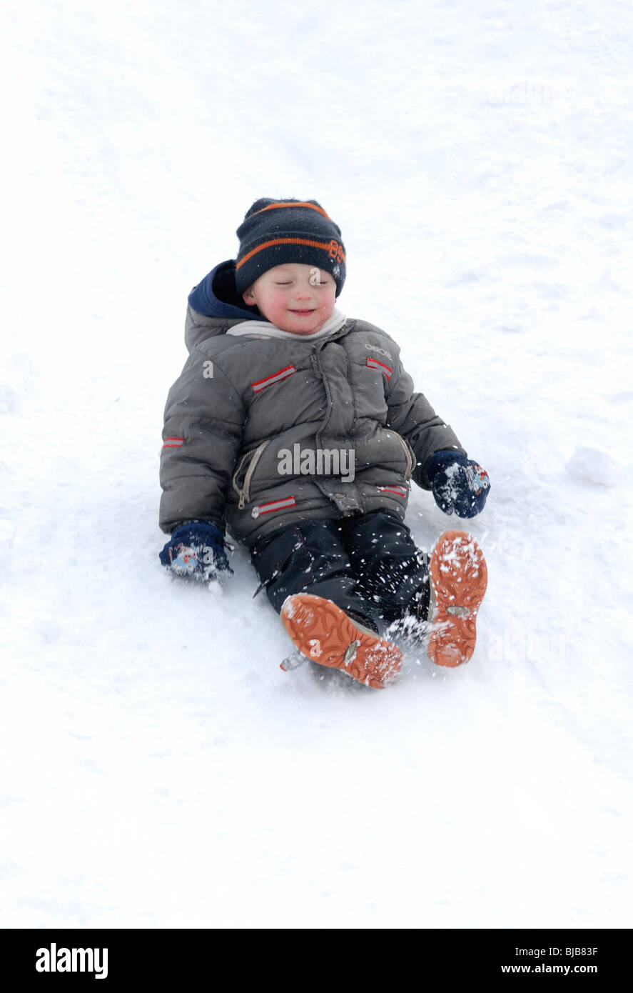 Lachendes Kind junge rutscht ein Schnee bedeckt Hill und spielen im Schnee Stockfoto
