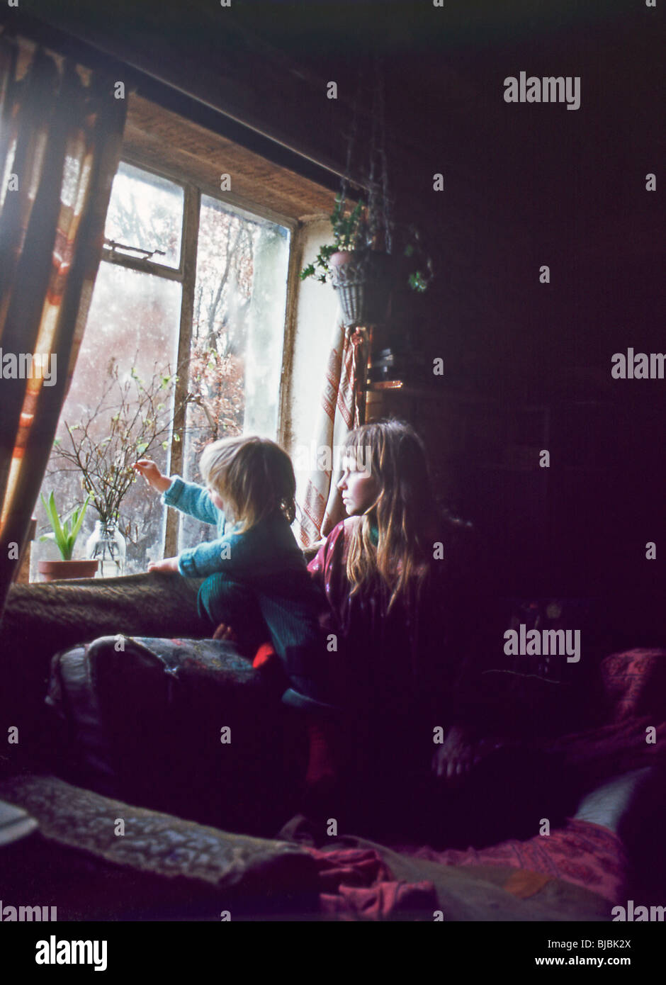 Hippie Mutter und Kind sitzen auf Sofa, spielen im Wohnzimmer und schauen aus dem ländlichen Häuschen Fensterhaus in den 1970er Jahren in Wales, Großbritannien KATHY DEWITT Stockfoto