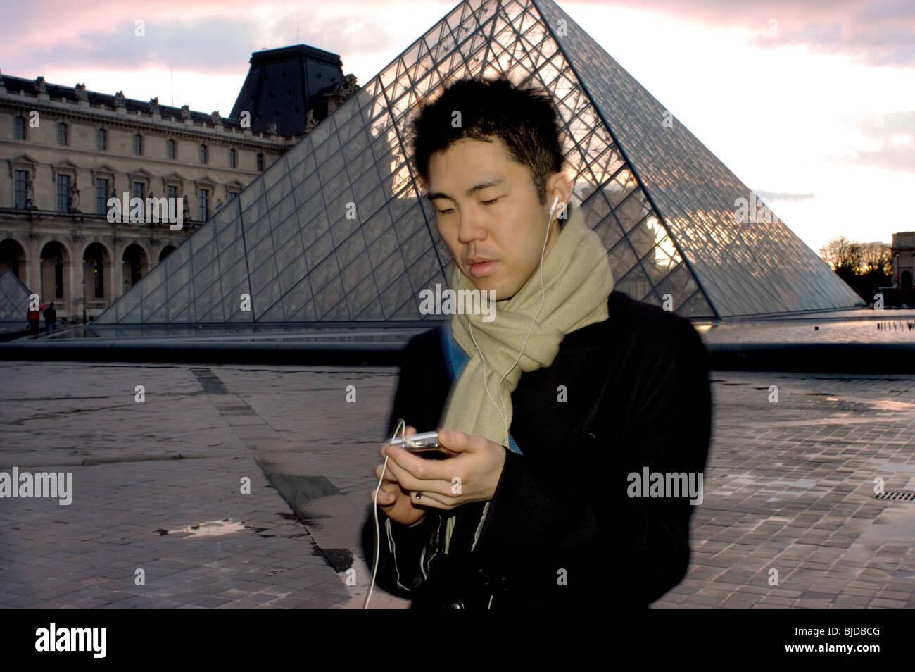 Paris, Frankreich - Portrait, Young man allein hört Nomad Device mit Ohrhörern in der Dämmerung, in der Nähe der Pyramide des Louvre Museum. Louvre, Leute im Freien Stockfoto