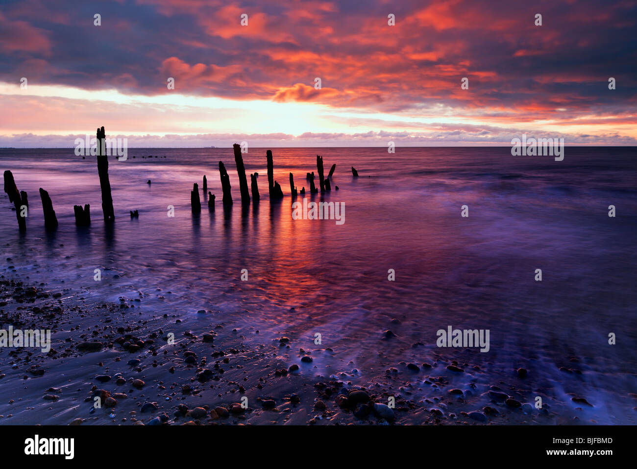 ein feuriger Sonnenaufgang spiegelt sich in den ausgehenden Strom mit verwitterten Buhnen im Vordergrund Winchelsea Beach Stockfoto