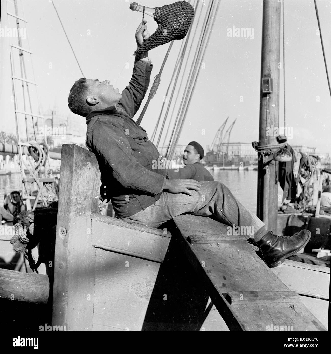 der 1950er Jahre in dieses historische Bild gießt ein Fischer sitzt am Boot an den Docks, Palma, Spanien, Wein seinen Mund Stockfoto