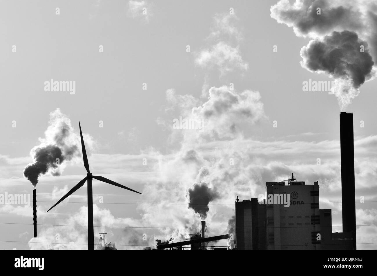 Schwarz-weiß-Bild von Rauch in den Himmel von einem Fabrikschlote als einer Windkraftanlage wogenden steht im Vordergrund Stockfoto