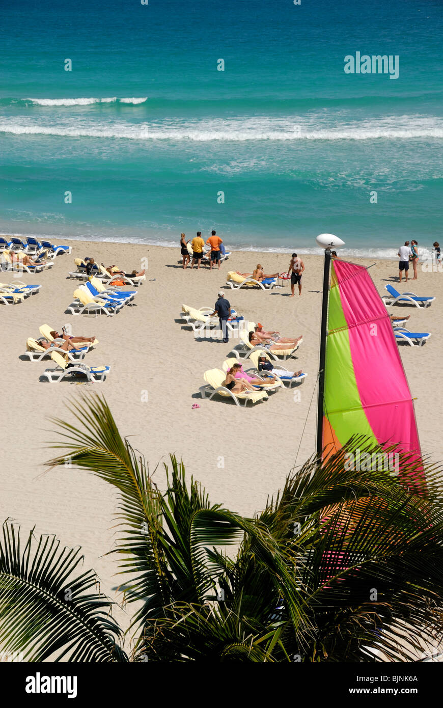 Sonnenanbeter in Liegestühlen am Strand von Florida, mit einem Segelboot im Vordergrund. Stockfoto