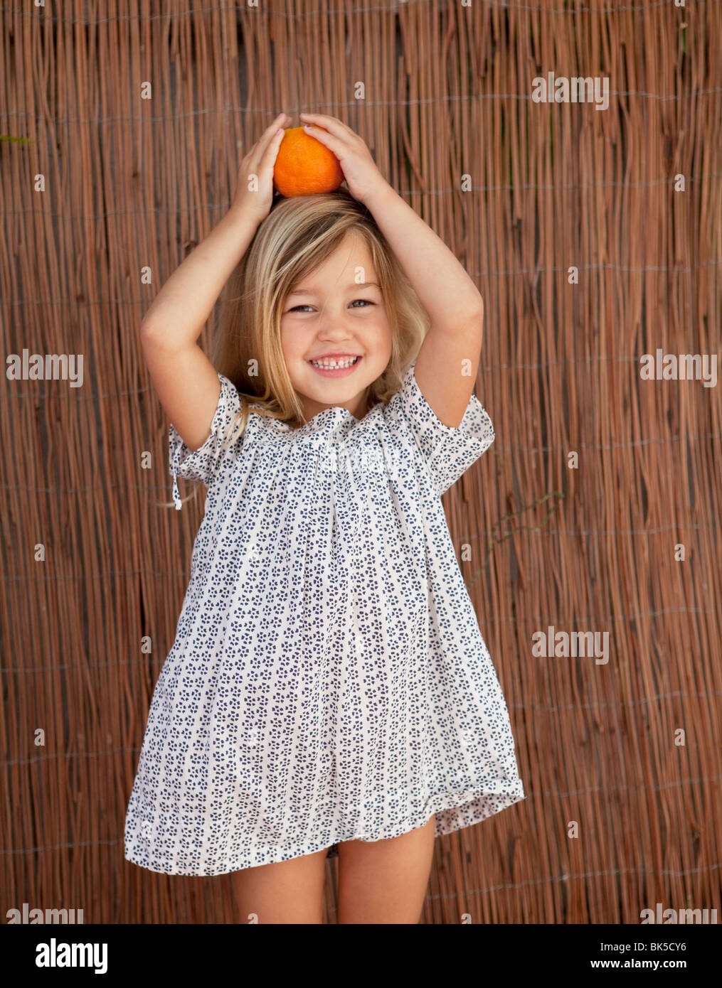 Junges Mädchen im Sonnenkleid hält eine Orange auf dem Kopf Stockfoto