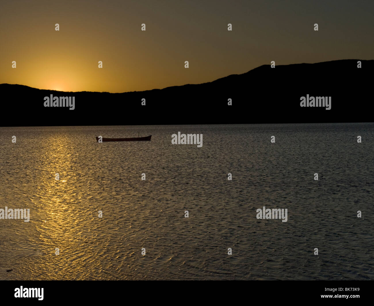 Über der Seeoberfläche, neben einem verlassenen Boot spiegelt sich die Sonne hinter den Bergen. Stockfoto