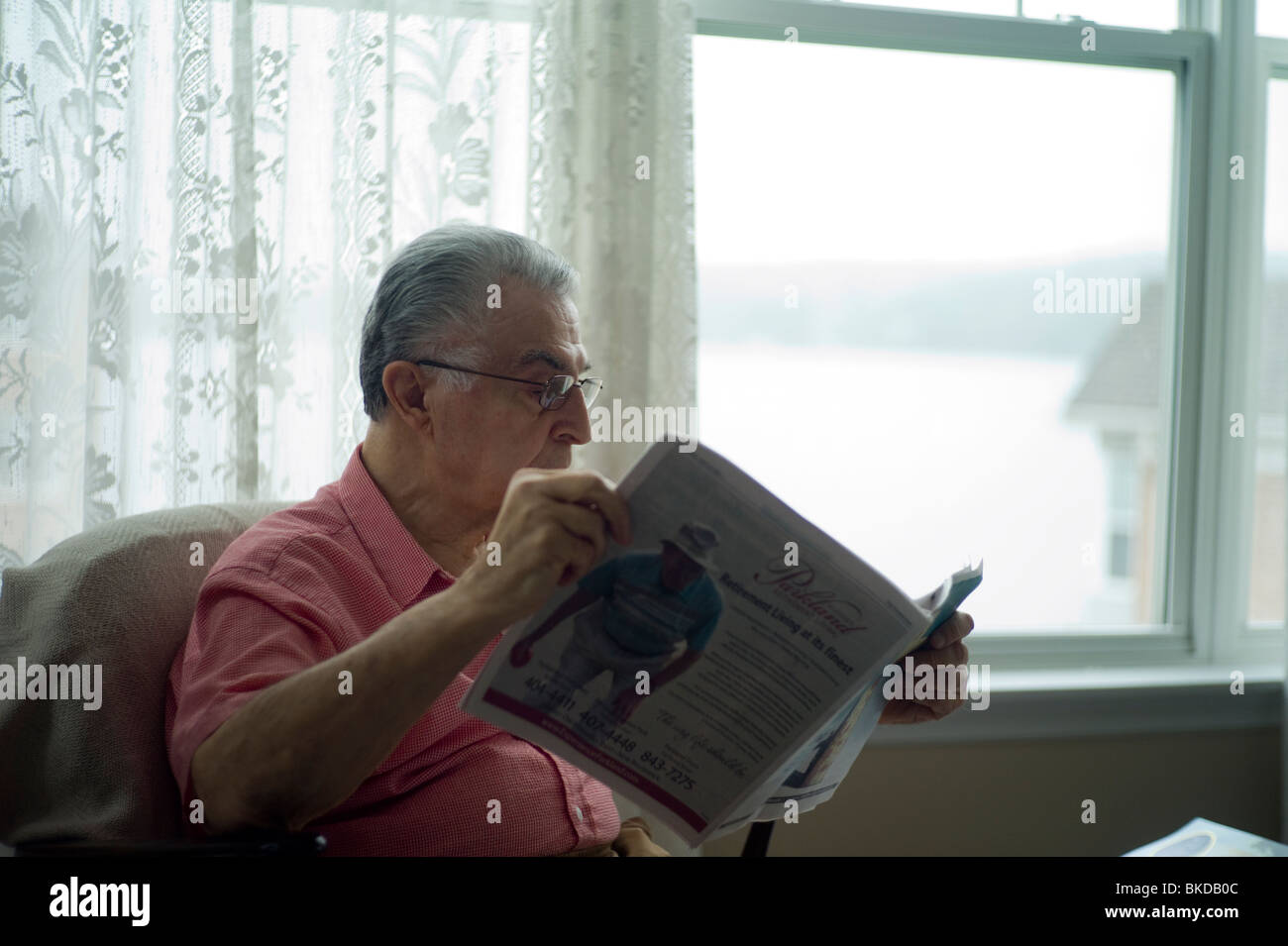 Alter Mann sitzt in Wohnzimmer Stuhl Fenster und Zeitung lesen. Stockfoto