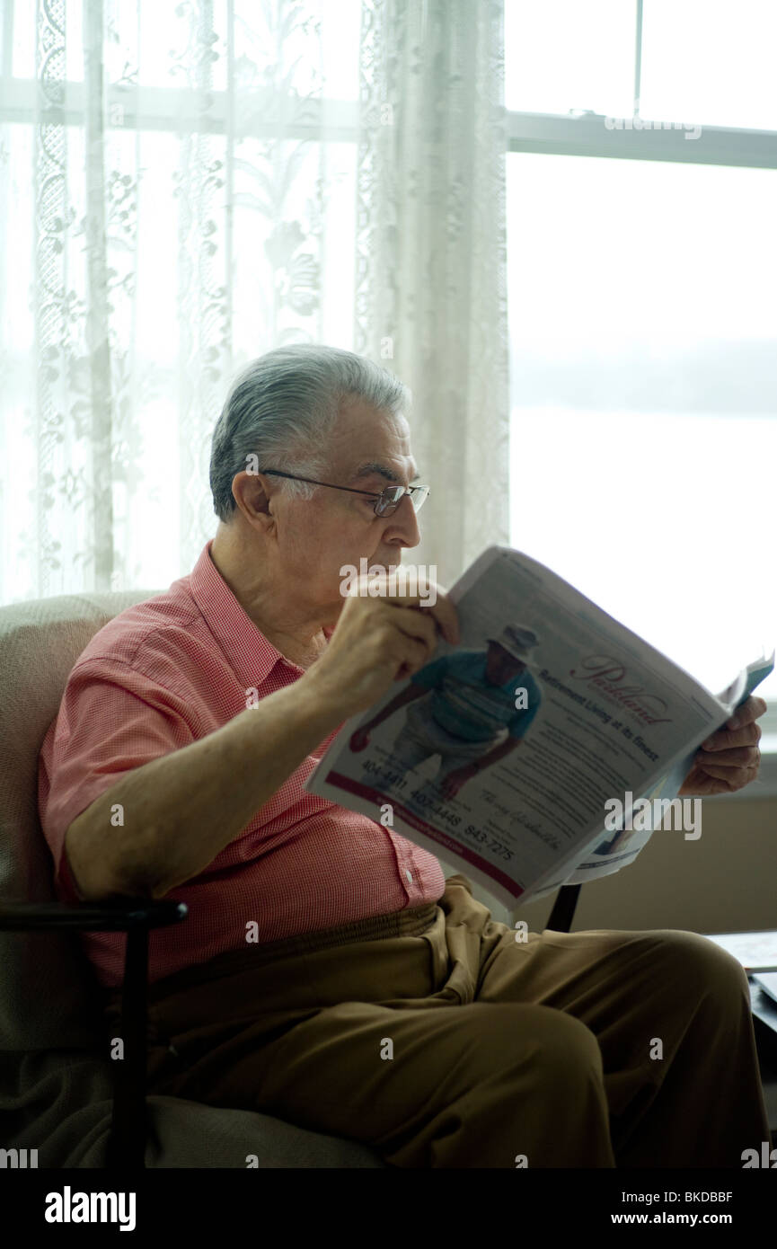 Alter Mann sitzt in Wohnzimmer Stuhl Fenster und Zeitung lesen. Stockfoto