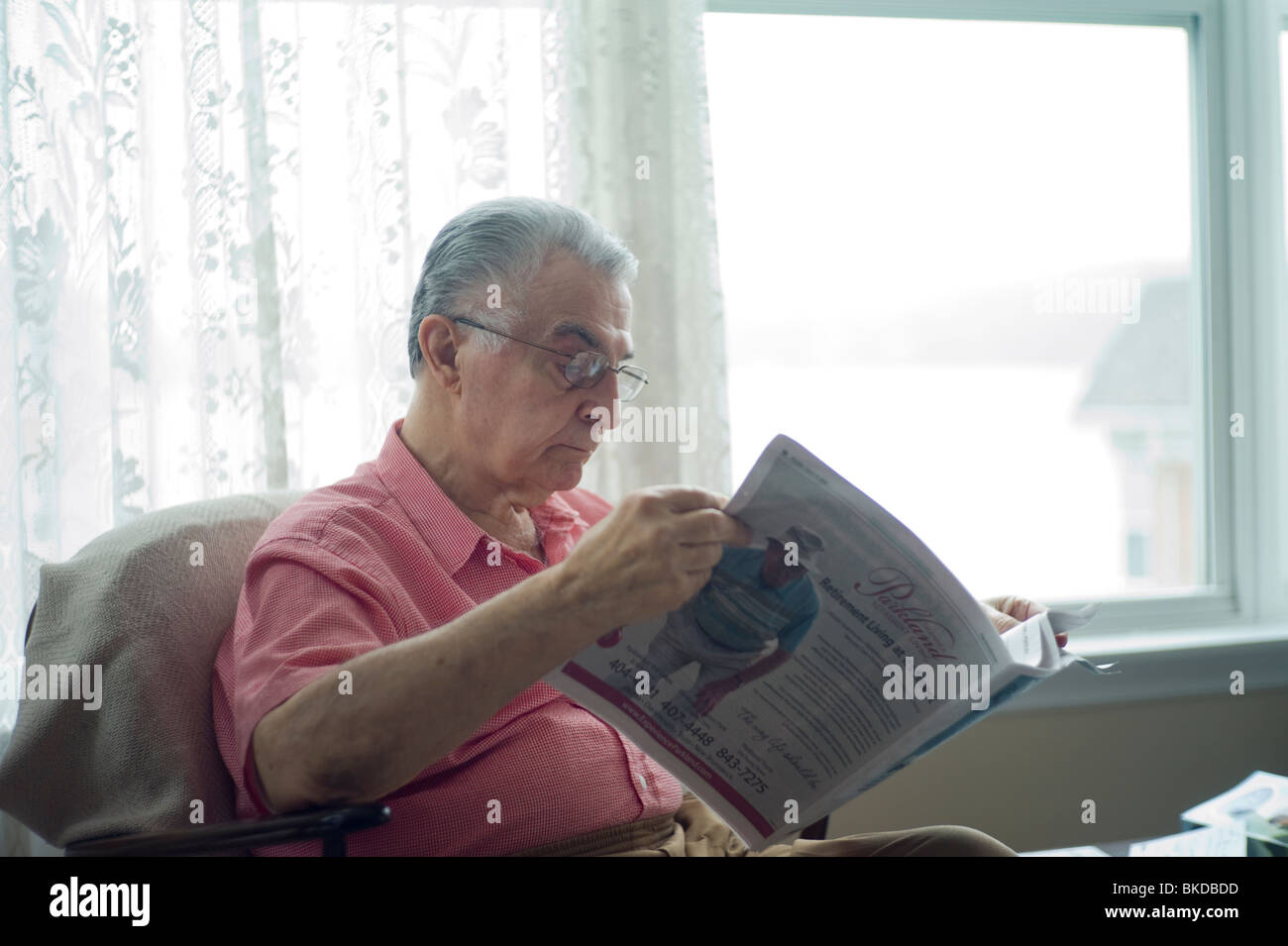 Alter Mann sitzt in Wohnzimmer Stuhl Fenster und Zeitung lesen. Stockfoto