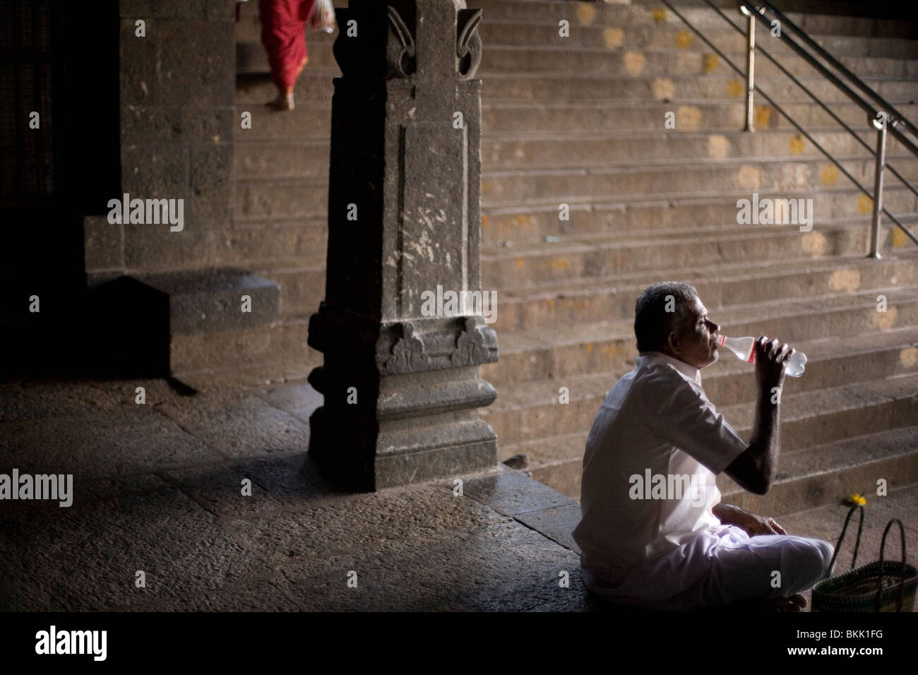 Ein Mann nimmt einen Schluck Wasser aus der Flasche, während seine Frau geht und ein Angebot in einem Schrein im Murugan Tempel in Swami macht Stockfoto