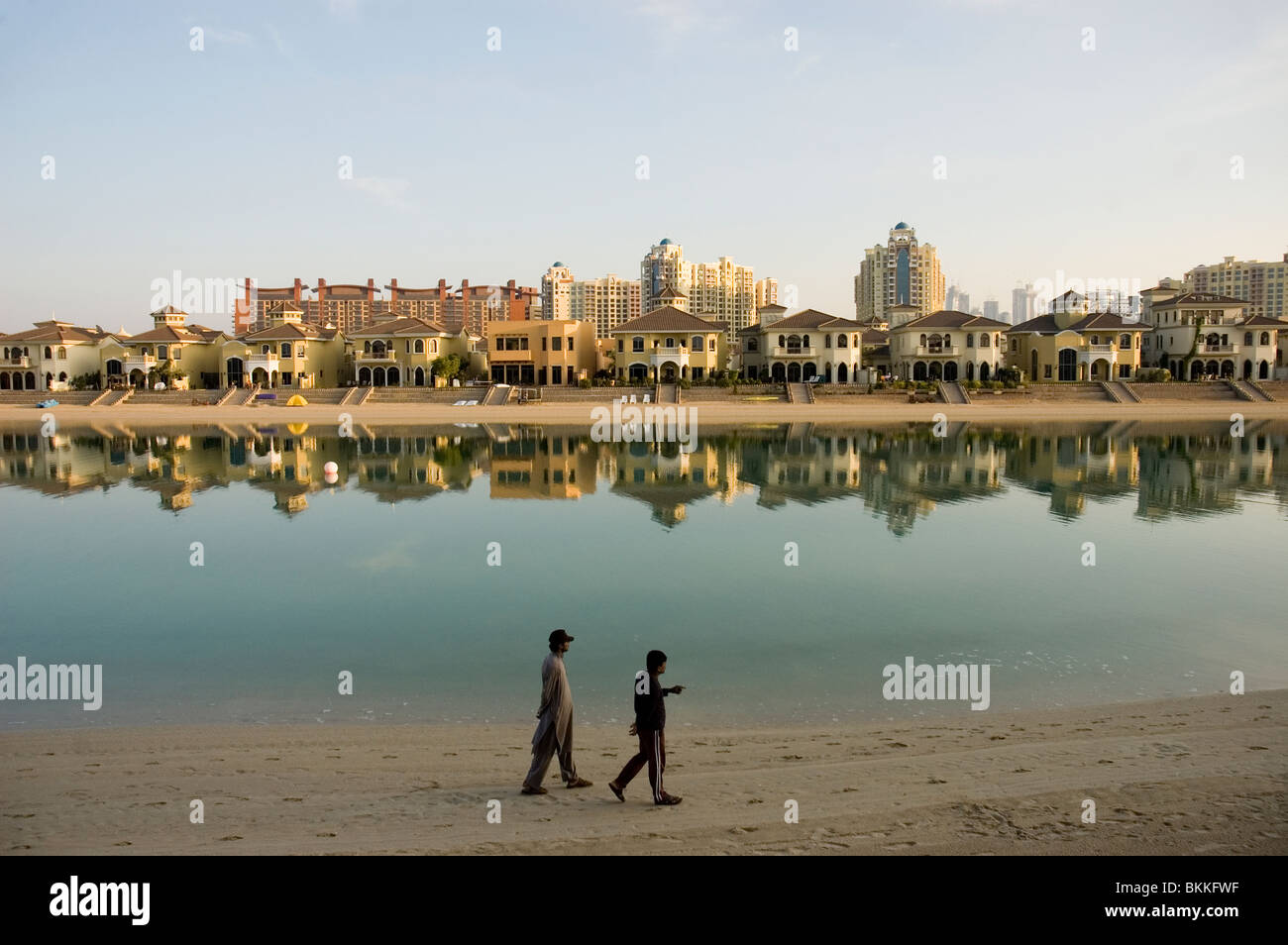 Zwei Männer zu Fuß am Strand entlang auf einem Wedel von der Palm Jumeirah mit Häusern auf der Handfläche und der Stadt Dubai im Hintergrund Stockfoto