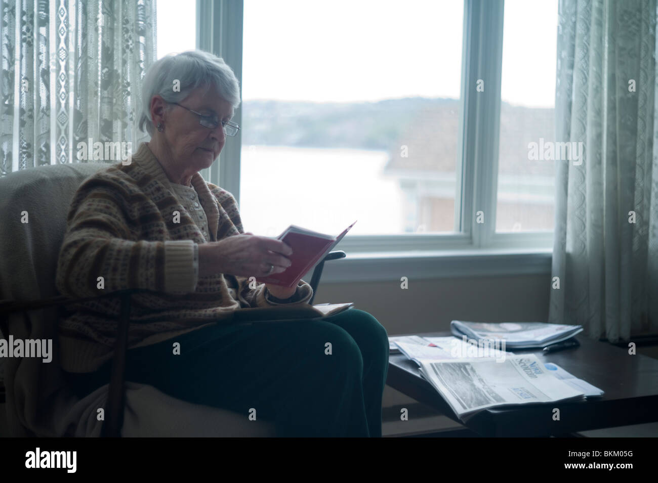 Alte Frau sitzt im Stuhl von Fenster und kleines Fotoalbum blättert. Stockfoto