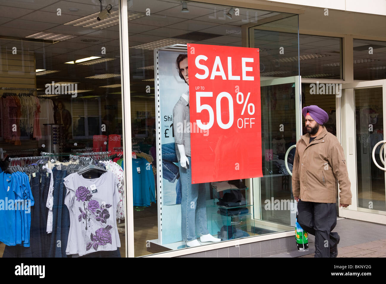 Ein Sikh mit einem lila Turban geht vorbei an einem Schaufenster anzeigen ein Verkaufsschild, Foto und Mannequin, Loughborough, England. Stockfoto