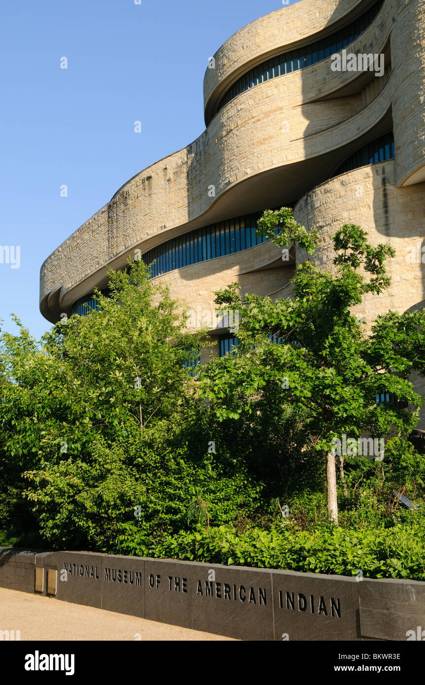 National Museum of the American Indian Washington DC // WASHINGTON DC – Außenansicht des National Museum of the American Indian der Smithsonian Institution, ein markantes, kurvenförmiges Sandsteingebäude. Üppiges grünes Laub verdeckt die Fassade teilweise, und der Name des Museums ist sichtbar, der in eine Steinmauer am Fuß geschnitzt ist. Stockfoto
