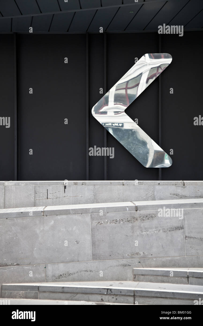 Reflektierende Metallpfeil gegen schwarze Wand mit Betonwand und Schritte im Vordergrund. Stockfoto