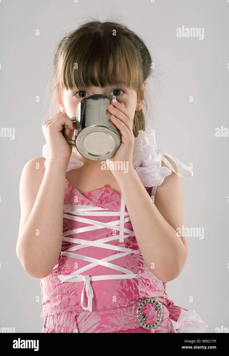 schöne Mädchen im rosa Kleid trinkt aus einem metallischen Becher auf grauem Hintergrund Stockfoto