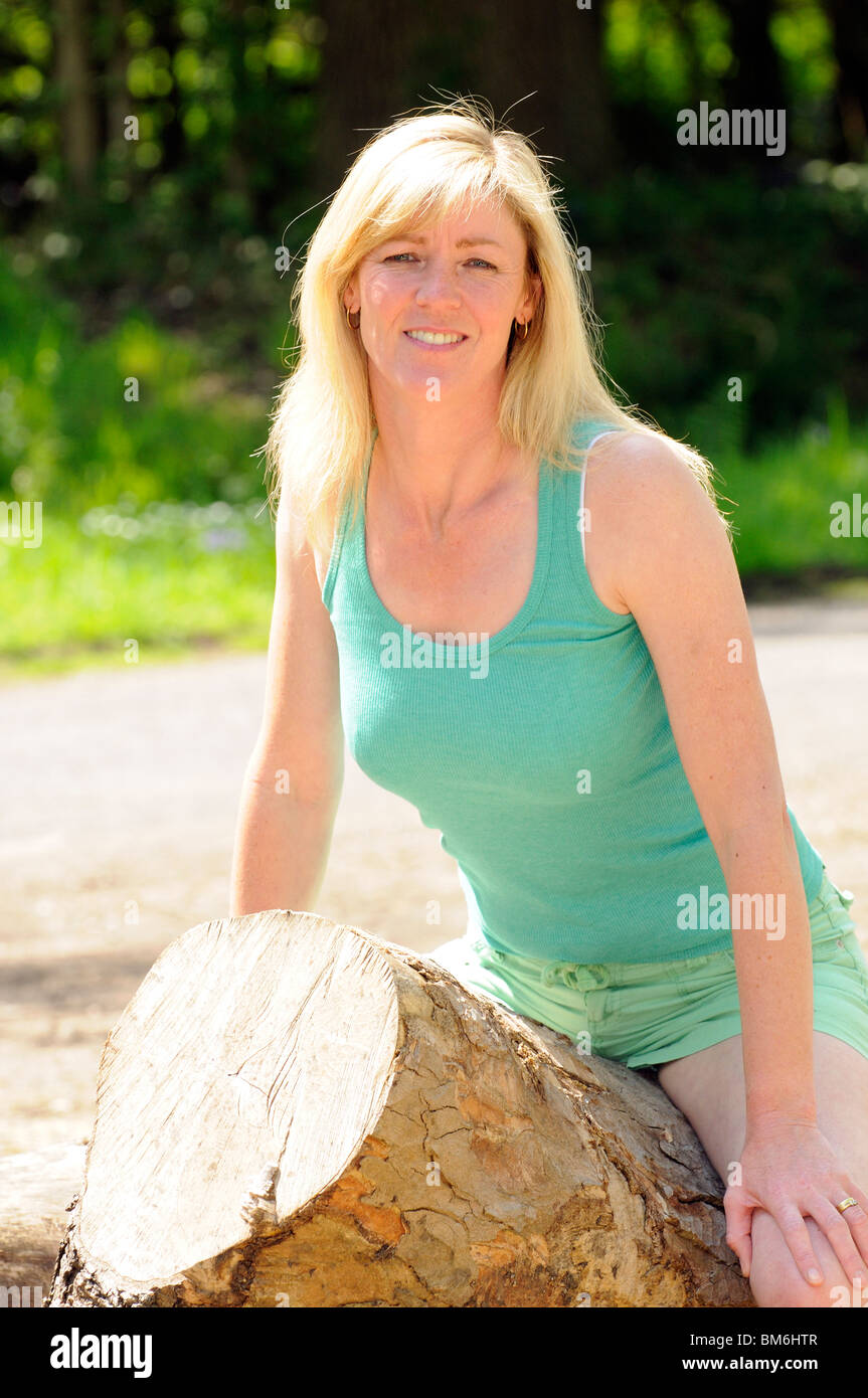 Porträt von Mitte vierzig blonde Frau im Freien sitzen auf einem gefällten Baum A Dreiviertel Körper Länge Ansicht Stockfoto