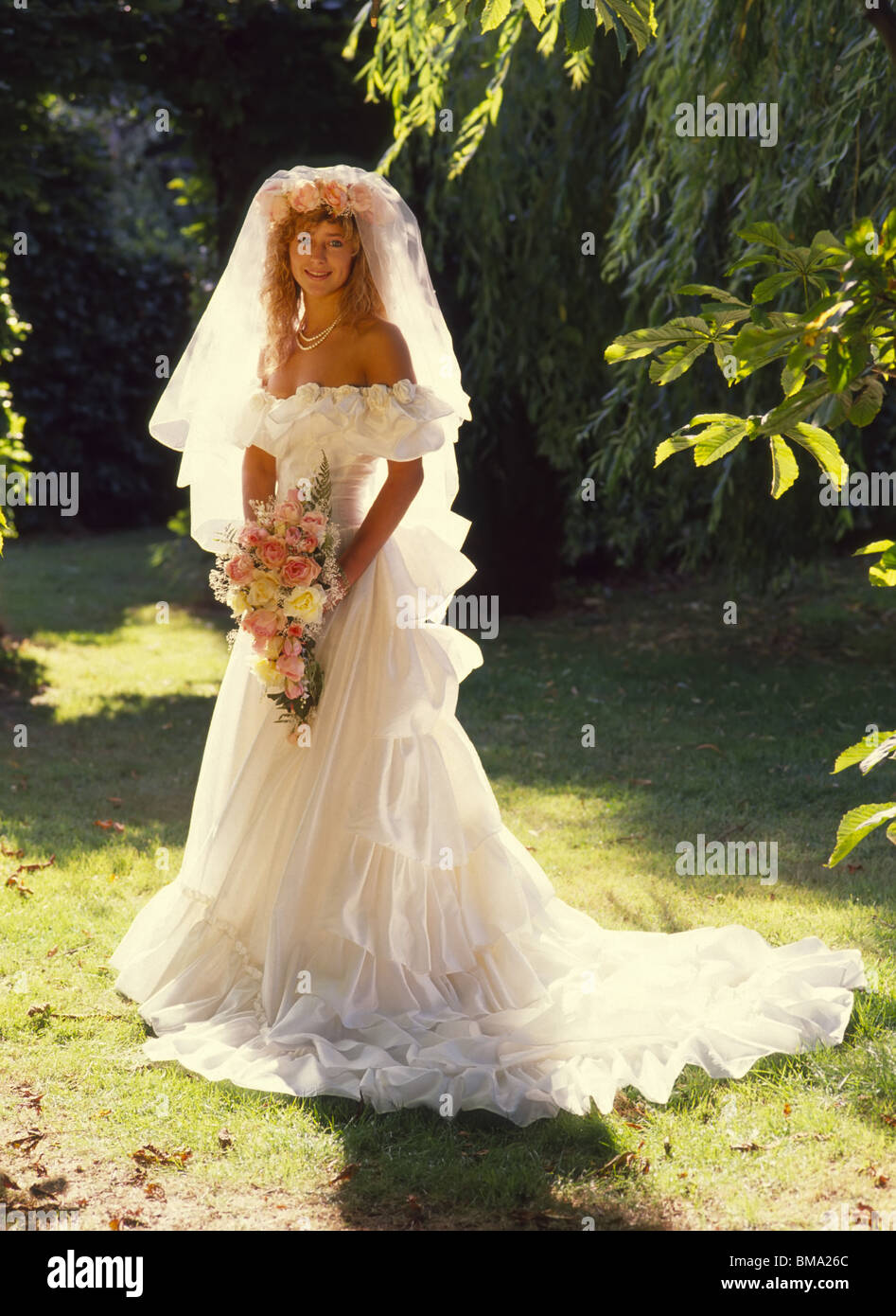 1989 hübsche junge Braut posiert für hinterleuchtete volle Länge von der Schulter Hochzeitskleid Foto mit Blumenstrauß 80er Jahre Modebild Essex England UK Stockfoto