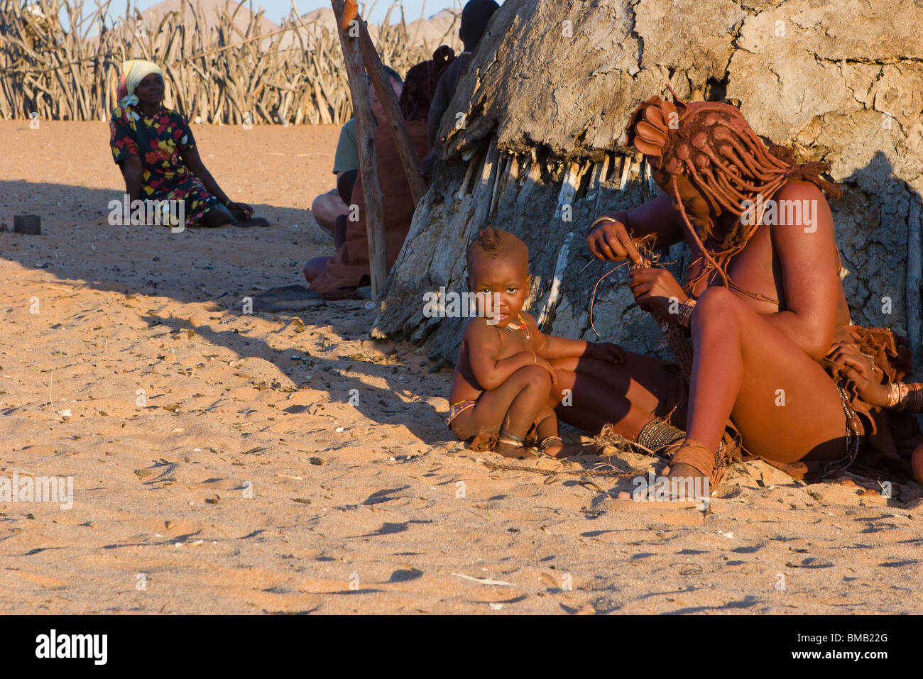 Nomadische Himba, Mutter und Kind das Weben in warmen Abend Sonne durch den Kuhmist Hütte in traditioneller Tracht und roter Ton Haare Stil gekleidet Stockfoto