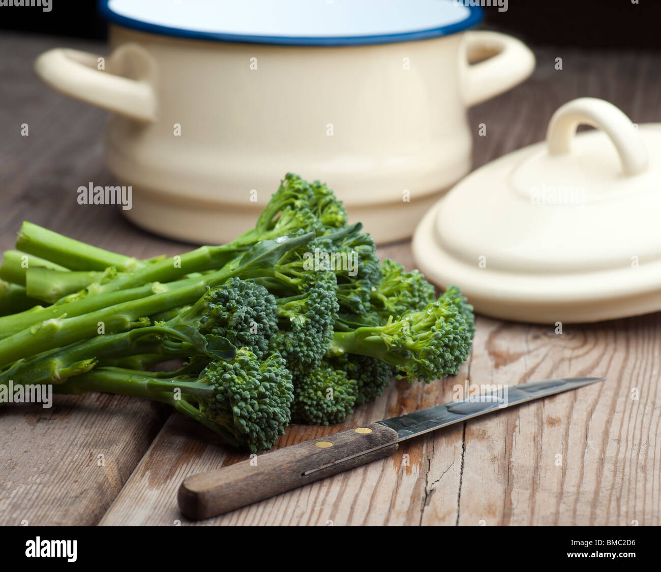 Frischer Brokkoli mit einem Gemüsemesser und eine Pfanne mit Deckel im Hintergrund auf ein Holz Küchentisch gelegt Stockfoto